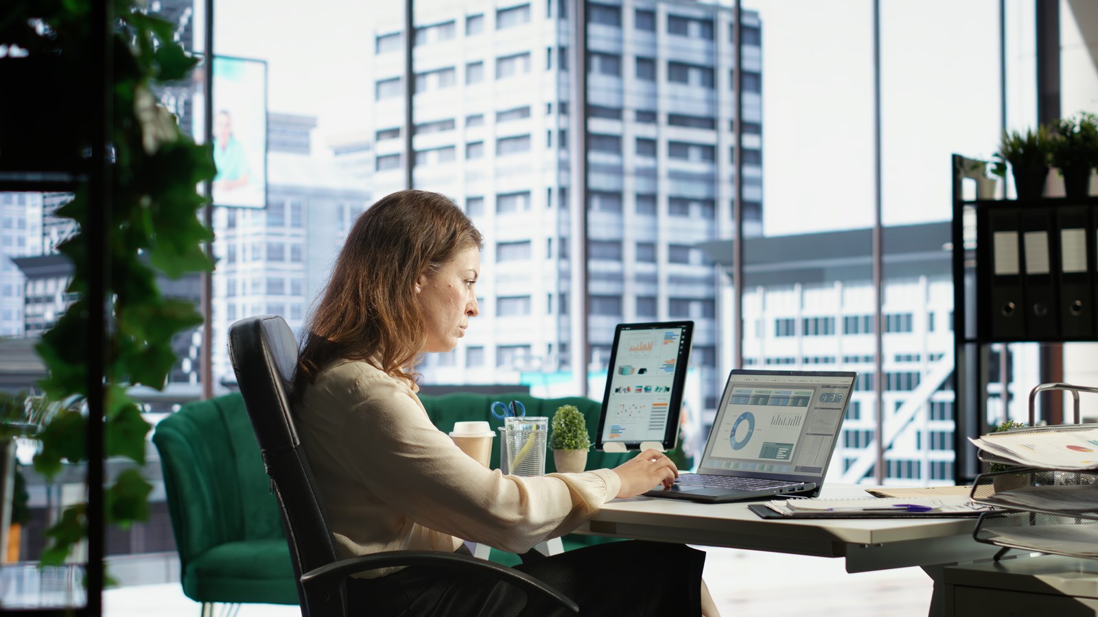Businesswoman working on a laptop and tablet at a desk beside a large city-view window