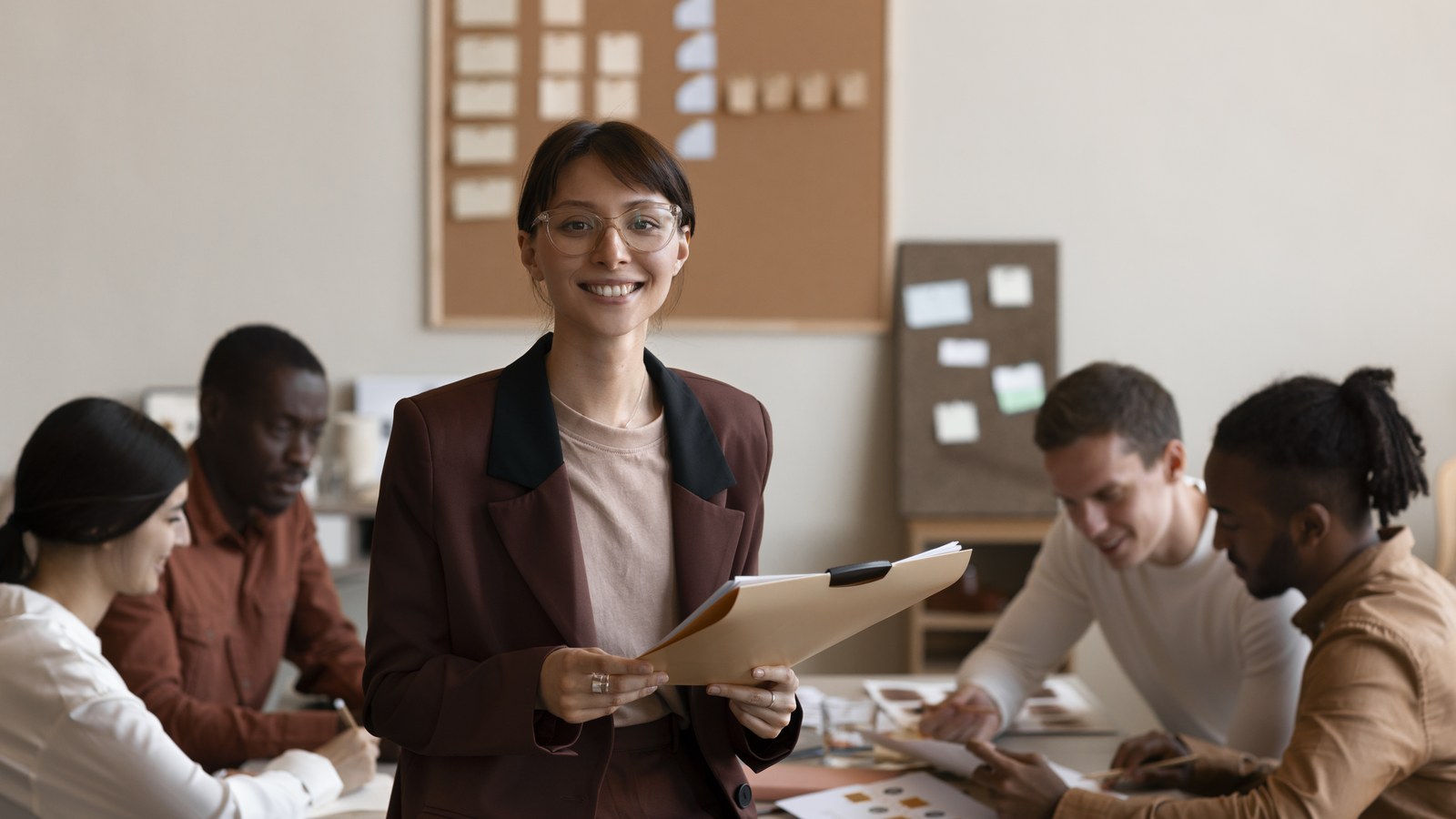Smiling manager holding a folder while team members work together at a table behind her
